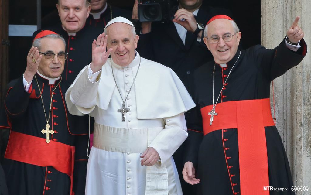 Pave Frans vinkar til folket iført kvit kappe og kvit kalott. Omgitt av kardinalar kledd i svarte kapper med raude band. Foto.