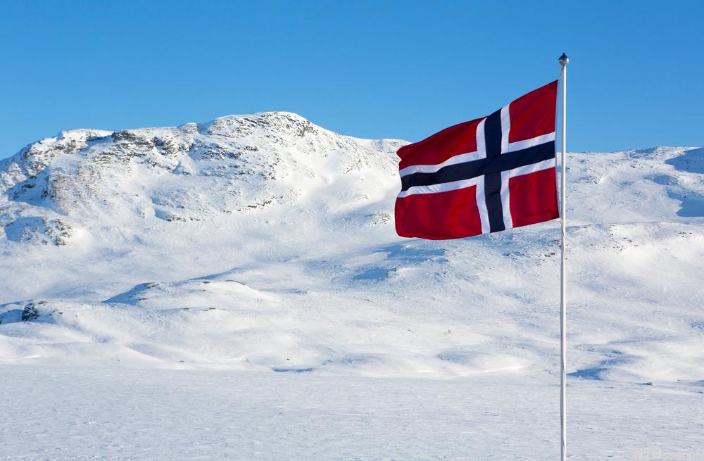 Norsk flagg vaier i vinden med snødekte fjell i bakgrunnen. Foto.