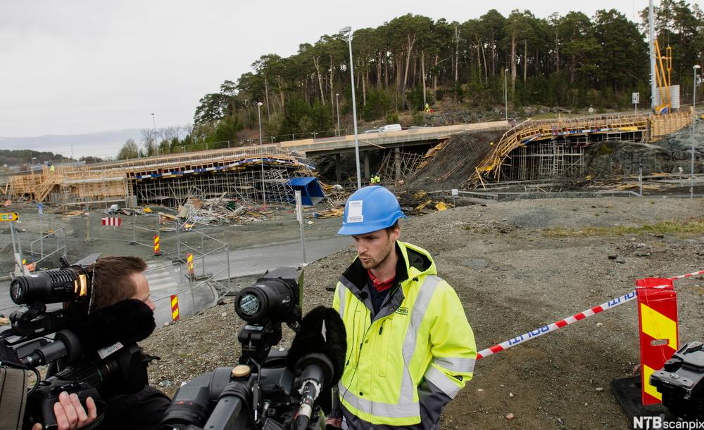 En mann med refleksvest og arbeidshjelm holder pressekonferanse på en byggeplass. I bakgrunnen ser det ut som en vei under oppføring har rast. Foto.