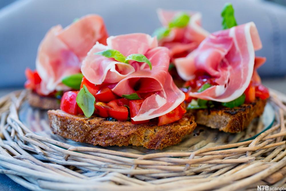 Tre bruschetta med spekeskinke og tomat, pyntet med basilikum. Foto.