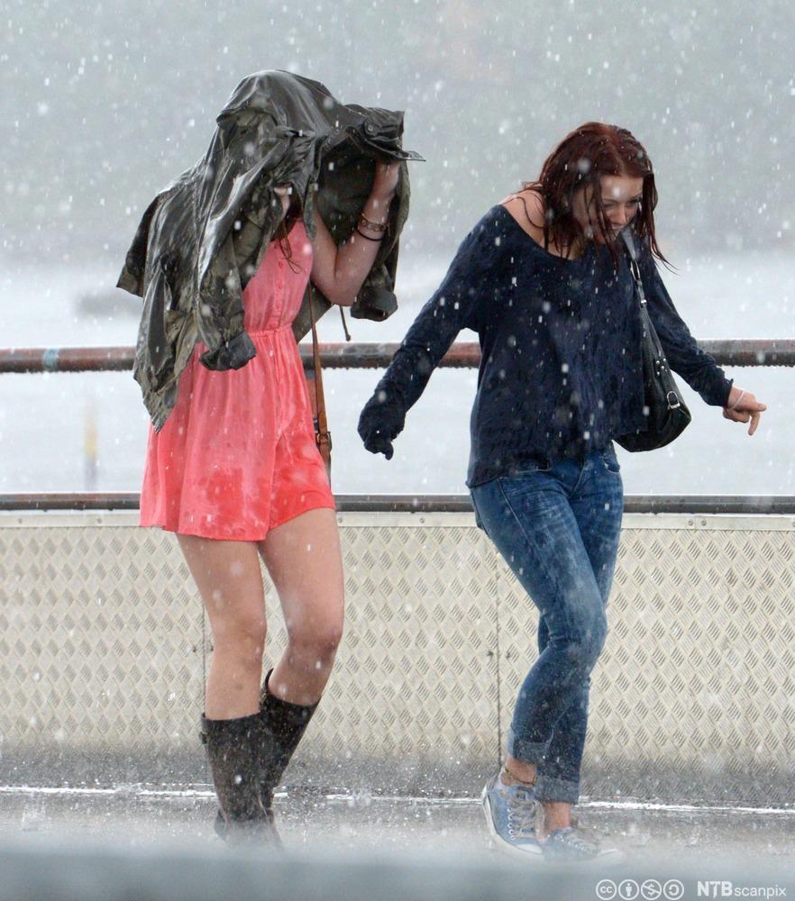 To jenter i regnvær. Den ene er kledd i en tynn kjole og holder ei jakke over seg som skjold mot regnet. Den andre har olabukse og genser og bøyer seg mot regnet. Foto.