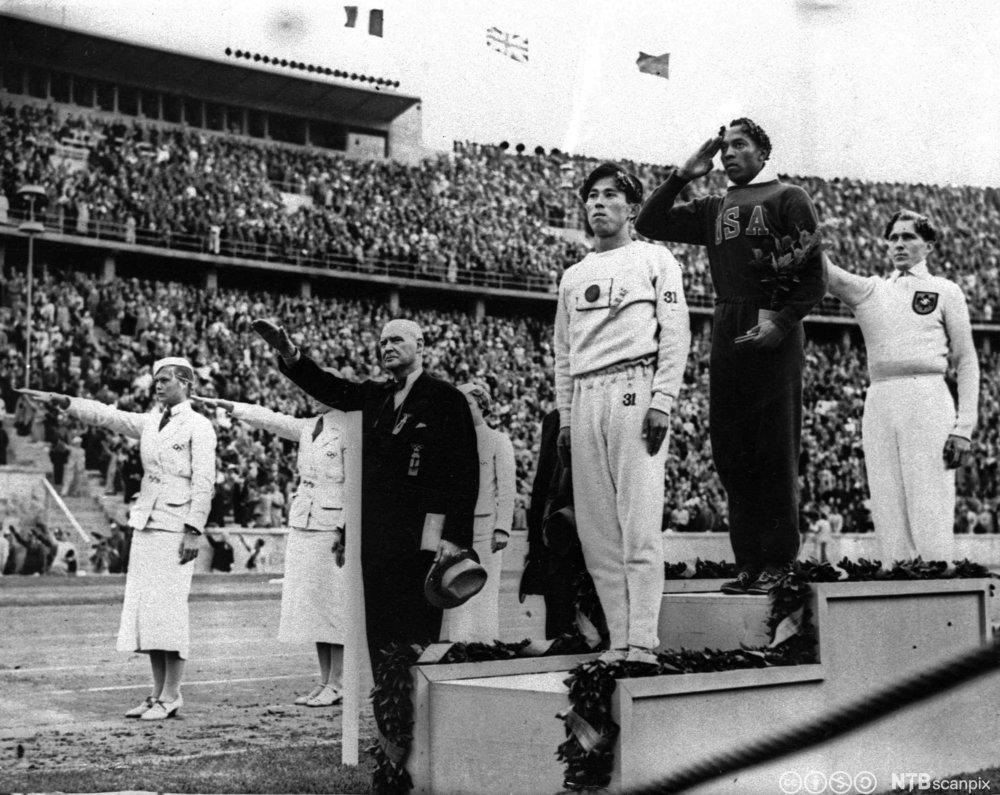 Den amerikanske gullmedaljevinneren Jesse Owens mottar  medaljen sin i Berlin-OL i 1936. Han står øverst på en seierspall med hånda opp mot pannen i en hilsen. Mannen på andreplass står med hendene rett ned. Den tredje på pallen og alle funksjonærene gjør nazihilsen. Svart/hvitt-foto. 