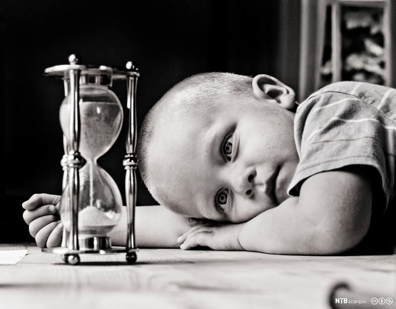 En liten gutt ligger og ser på et timeglass. Foto.