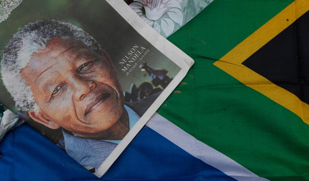 Ei avisframside med eit stort bilete av Nelson Mandela er lagd på eit sørafrikansk flagg. Avisa har Mandelas namn, fødsels- og dødsdato som påskrift. Foto.