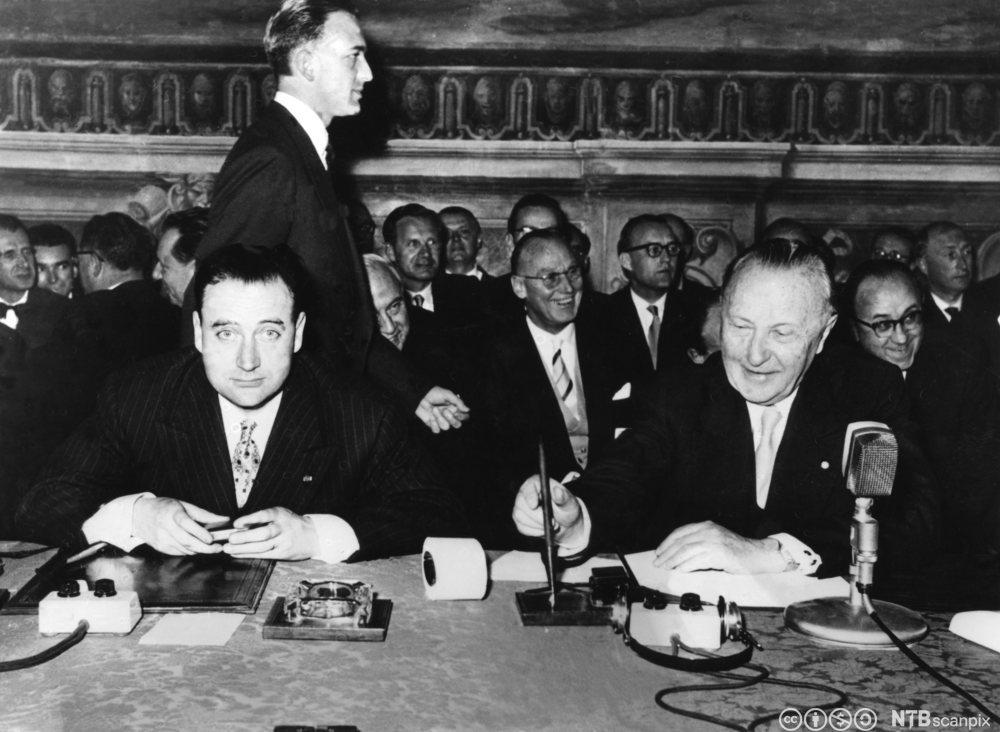 To menn i dress og slips sitter ved et bord. Den ene mannen har en bunke papirer foran seg og tar tak i en penn. I bakgrunnen er det mange menn. Flere er tydelig i godt humør. Svart-hvitt foto. 