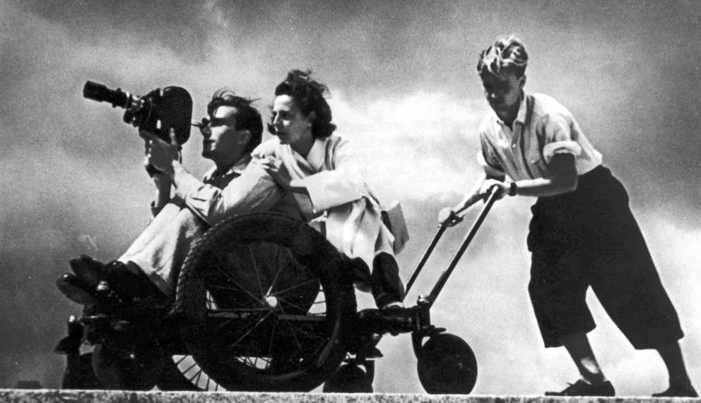 Foto i svart-kvitt av ein kameramann og ei kvinne som begge sit oppi ei vogn medan dei filmar. Vogna blir dytta av ein ung mann. Alle tre er kledde i 30-talsklede.