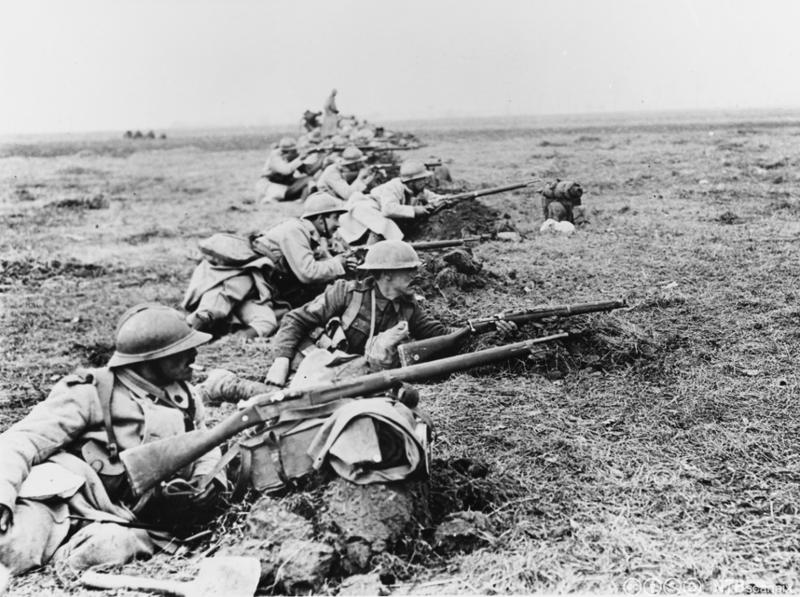 Bilde av soldater som ligger i stilling på ei lang rekke i ei grøft i et flatt landskap. De har geværer og hjelmer. Bildet er av britiske og franske soldater som dekker en vei i slaget ved Somme. Foto.