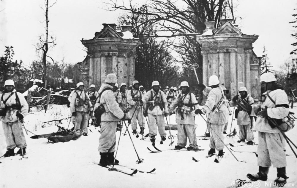 Hvitkledde soldater på ski står foran en portal eller lignende i klassisk arkitektur. I bakgrunnen en allé av trær. Foto. 