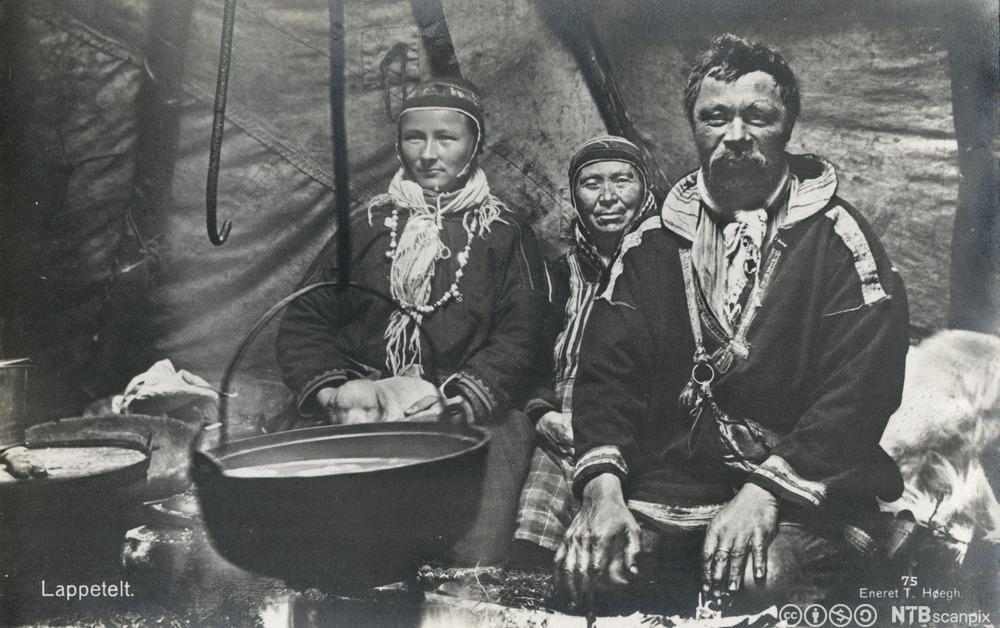 En samisk familie på 1930-tallet. Familiemedlemmene har på samedrakter og sitter inne i et telt. Foran henger ei gryte. Foto. 