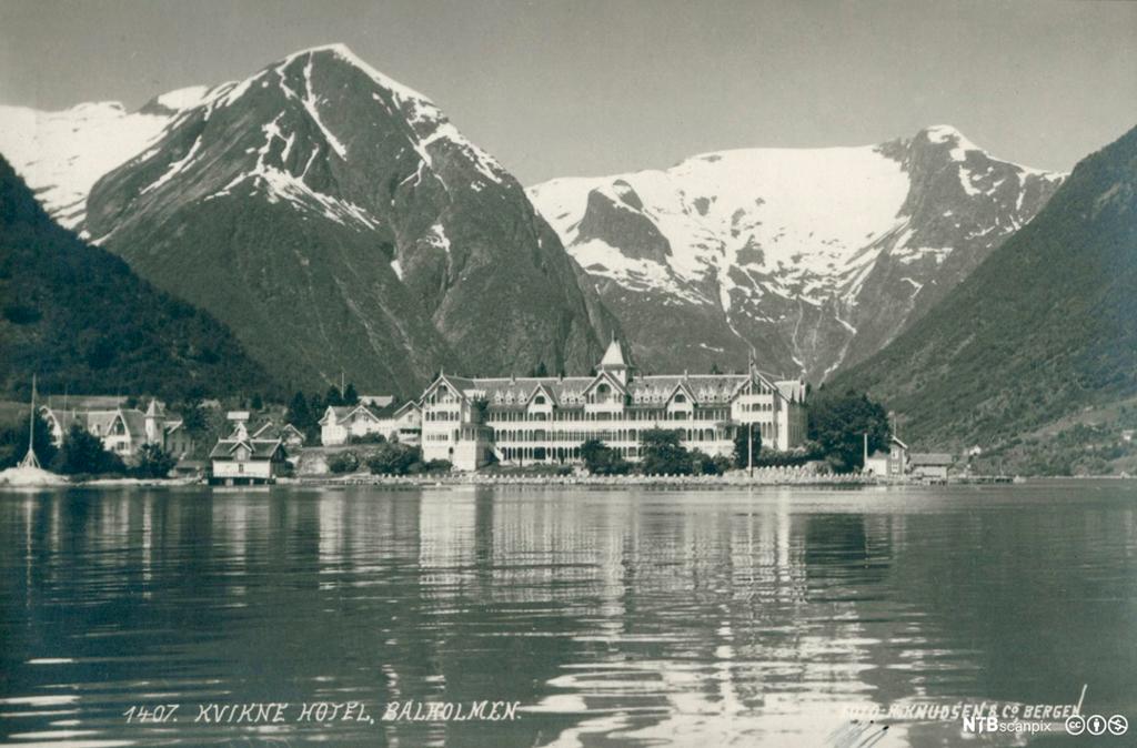 Utsikt over en fjord mot Kvikne Hotell, Balestrand med fjell som stiger opp bak. Foto.