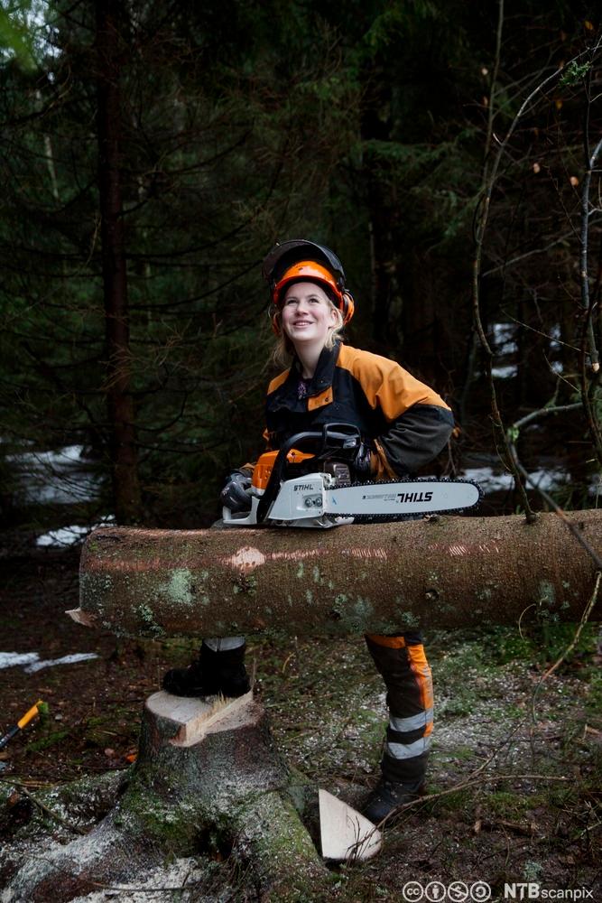 A forrest worker is standing by a tree, holding a chain saw. She's dressed in working clothes and a protective helmet. Photo.