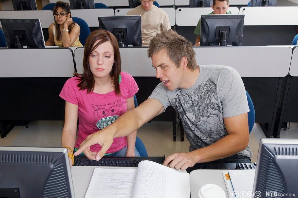 To studenter i et klasserom med bøker og datamaskin. Foto.