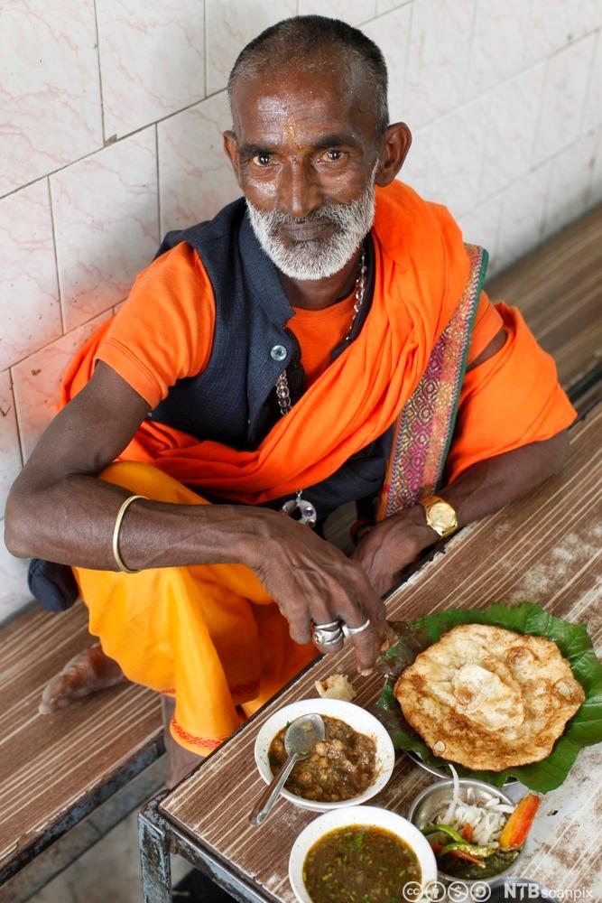 En sadhu spiser vegetarmat på en restaurant. Han har fargerike klær, gullklokke og sølvringer. Foto.