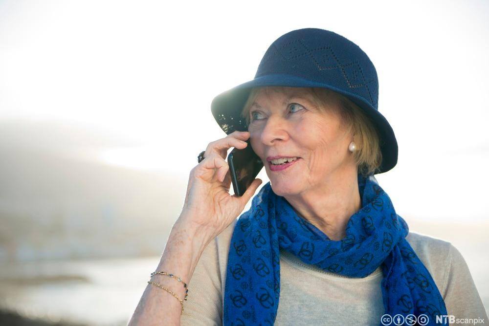 Eldre kvinne med hatt og skjerf snakker i mobiltelefonen. Foto.