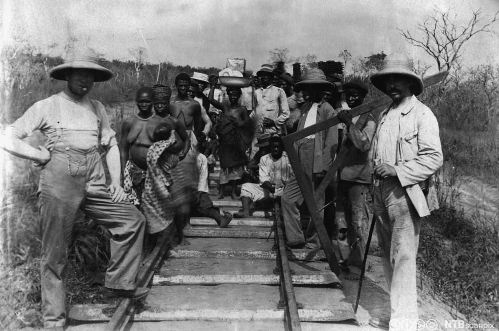 Både menn og kvinner i ulike klede står ved eit jernbanespor. Foto. 