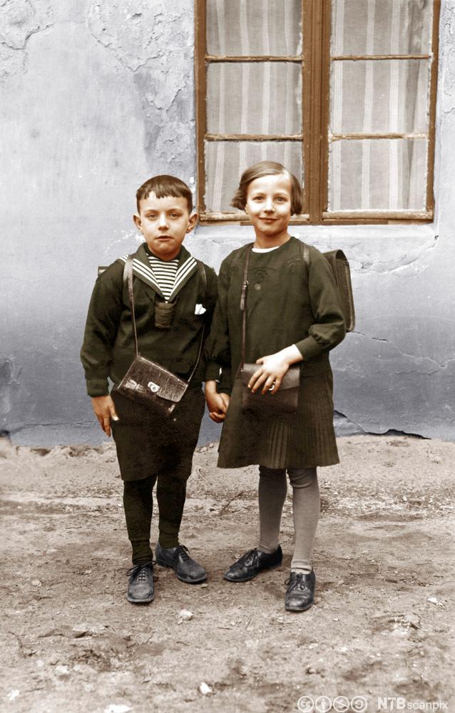 To skolebarn i småskolealder, gutt og jente. De holder hverandre i handa. Begge bærer en ransel på ryggen og ei lita veske med lang skulderreim. Tysk foto fra 1920-åra.