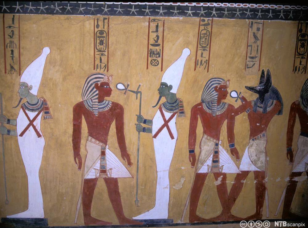 Seks figurer malt på en vegg i gammel egyptisk stil. To av dem bærer faraoens hodeplagg og to av dem er kledd i hvitt og bærer en stav. Den siste figuren er guden Anubis med menneskekropp og hodet til en sjakal. Foto.