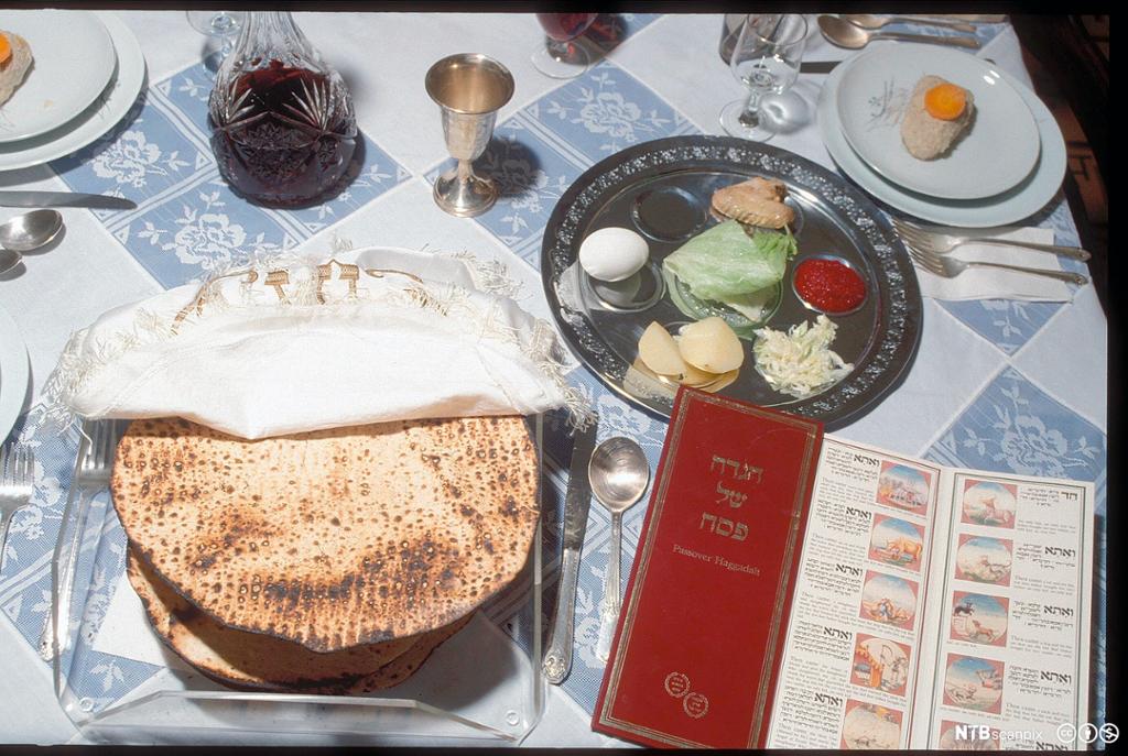 Bilde som viser de symbolske rettene i et jødisk seder-måltid. En bunke flatbrød, et glass rødvin og tallerken med ulike matvarer er oppstilt på et bord. Foto.