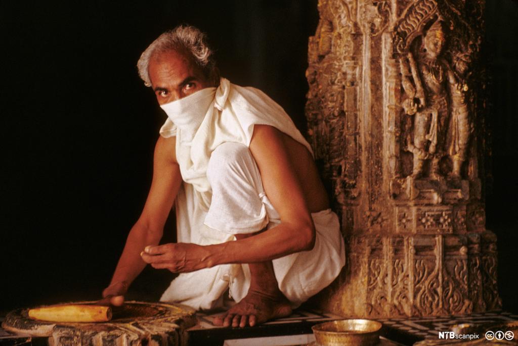 Mann med hvitt tørkle over munn og nese sitter på et gulv med en søyle bak seg. Han holder i noe laget av tremateriale. Foto.