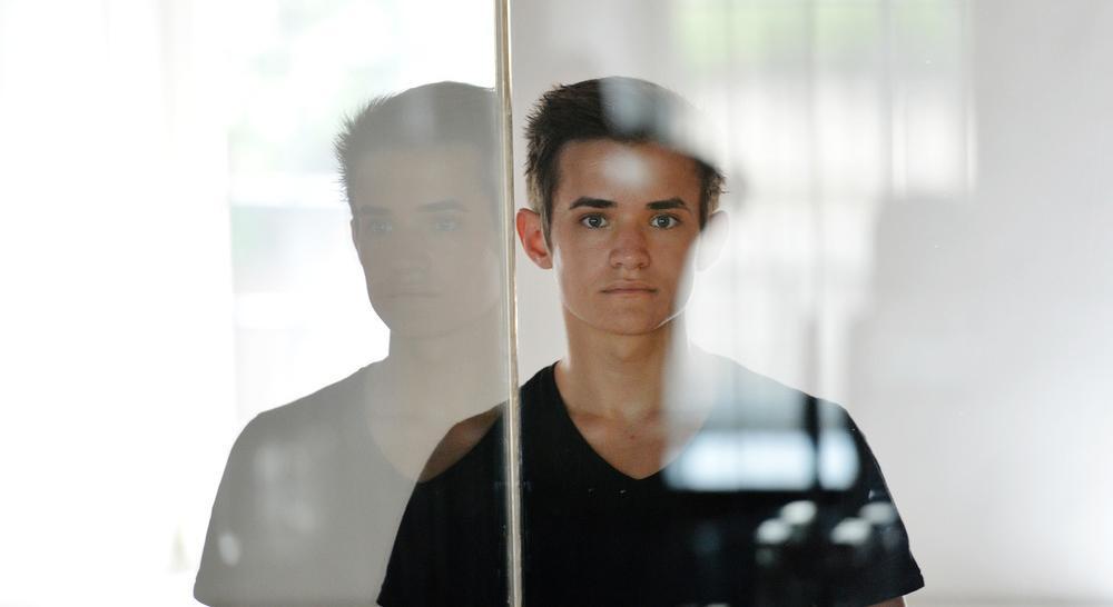 Ein ung gut ser rett inn i kameraet. Han står ved eit vindauge slik at vi ser refleksjonen av han til venstre. Foto.