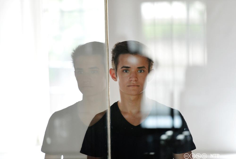 En ung gutt ser rett inn i kameraet. Han står ved et vindu slik at vi ser refleksjonen av han til venstre. Foto.