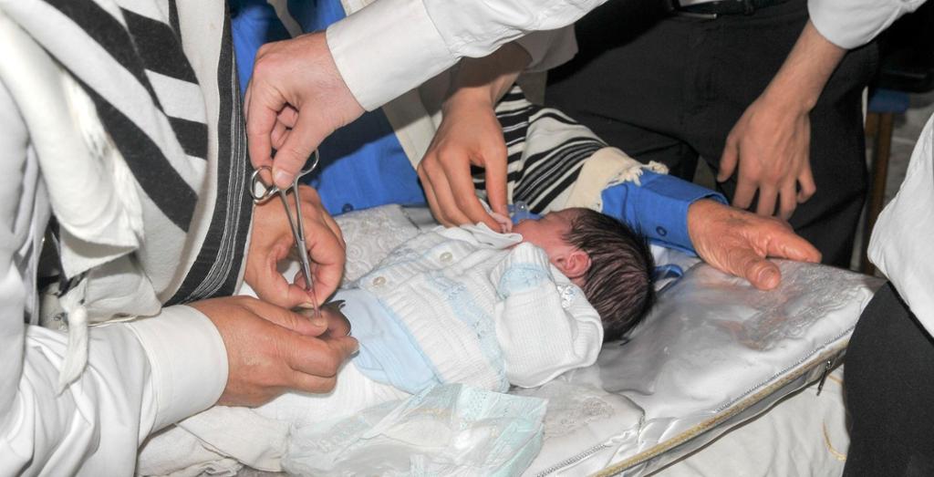 En guttebaby ligger på et staselig underlag og blir omskåret. Flere personer står omkring; en holder en saks. Foto.