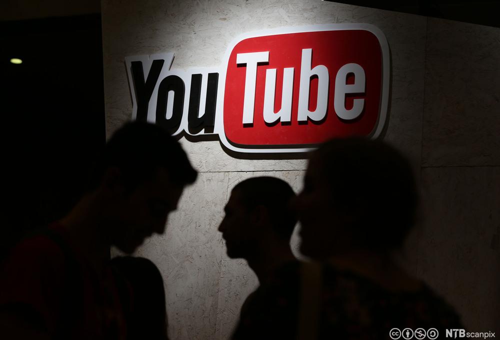 YouTube-logoen på ein vegg, tre silhuettar av personar under. Foto.