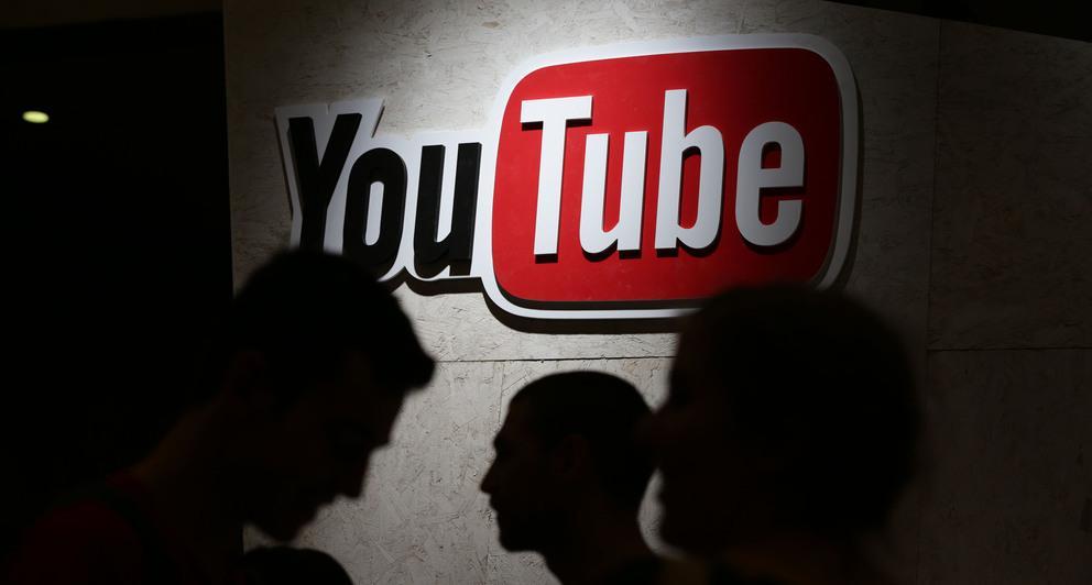 Fotografi av ein stor YouTube-logo på ein opplyst vegg med skuggar av menneske i framgrunnen.