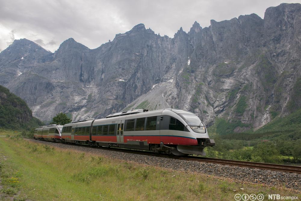 NSB-tog som køyrer på togskinner i natur med grøne sletter og fjelltoppar i bakgrunnen. Foto. 