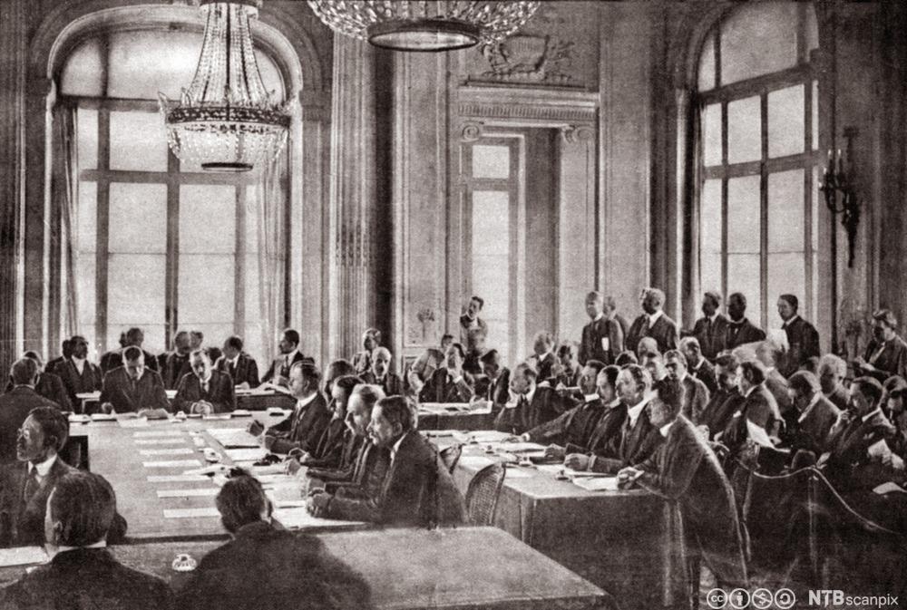 Bildet viser mange menn som sitter ved store bord i et stort rom. Bak i rommet sitter og står det enda flere menn. Det er høye vinduer og lysekroner i taket. 