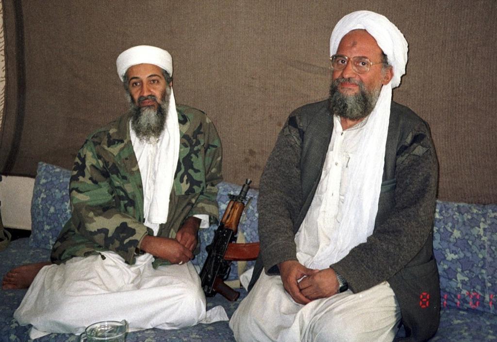 To eldre menn med skjegg, hvite kjortler, kamuflasjefargede jakker  og hvite hodeplagg sitter ved siden av hverandre på gulvet. Mellom dem ligger et maskingevær. Foto.