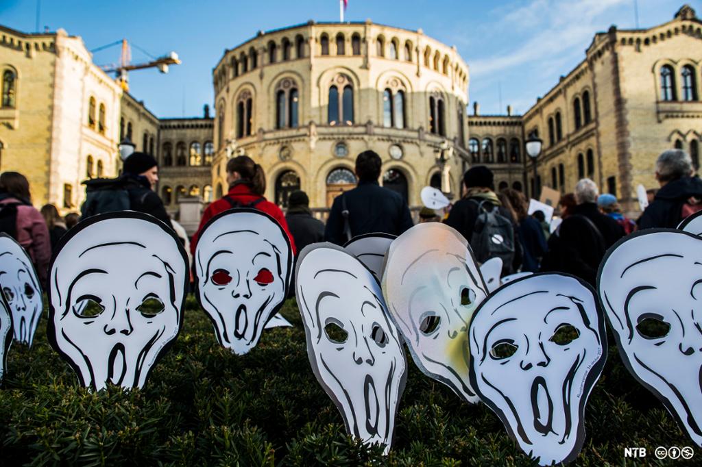Ei grupper personer demonstrerer foran Stortinget. På plenen bak dem står mange masker formet som en svart-hvit utgave av motivet i maleriet Skrik. Foto.