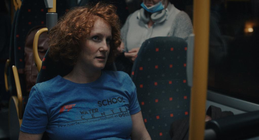 En ung kvinne med rødt hår og blå genser sitter på en buss. Hun har et skeptisk uttrykk. Foto.
