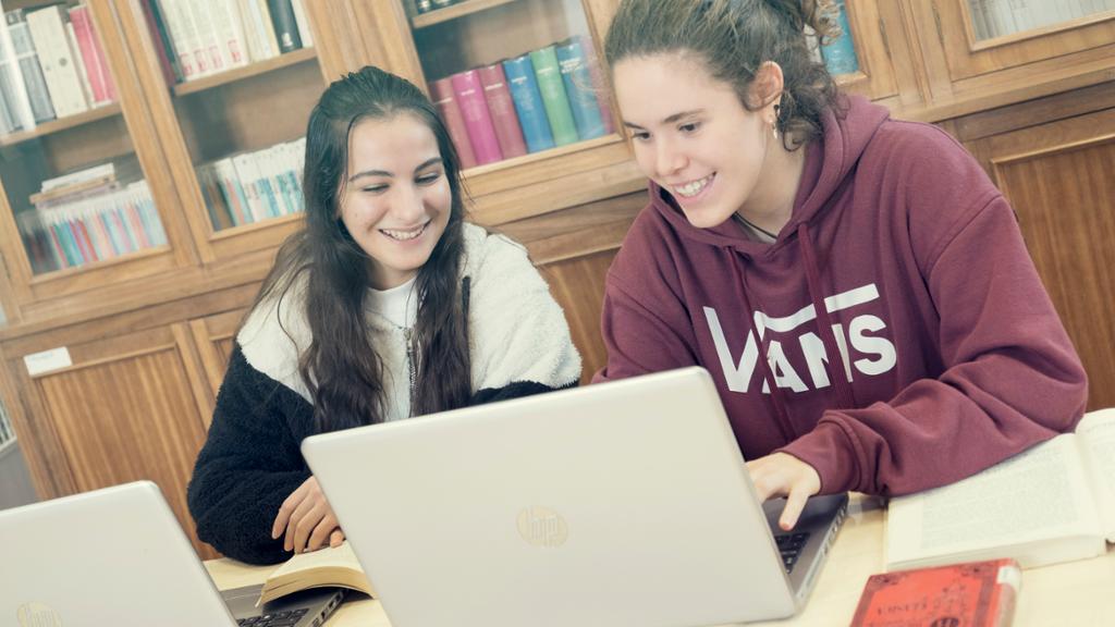 To smilende jenter studerer sammen med laptop og bøker i et bibliotek. Foto.