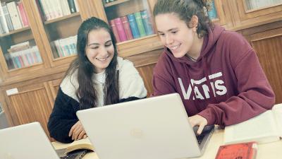 To smilende jenter med hver sin laptop samarbeider på et skolebibliotek. Foto.