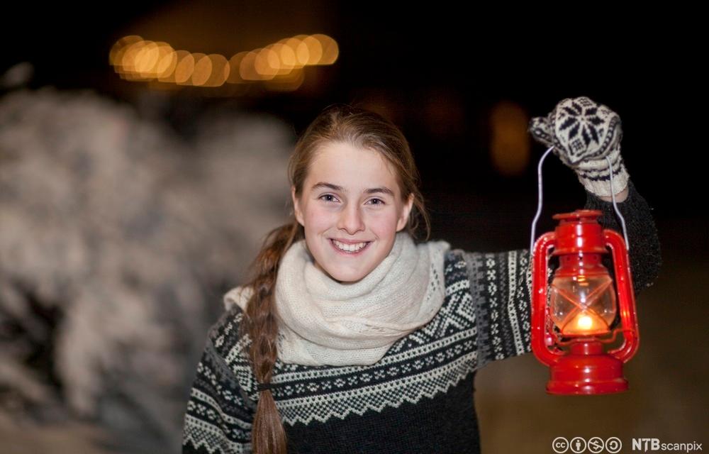 Blid jente med flette, som har på seg strikkegenser, strikkesjal og selbuvottar, held ei raud lykt. Foto.