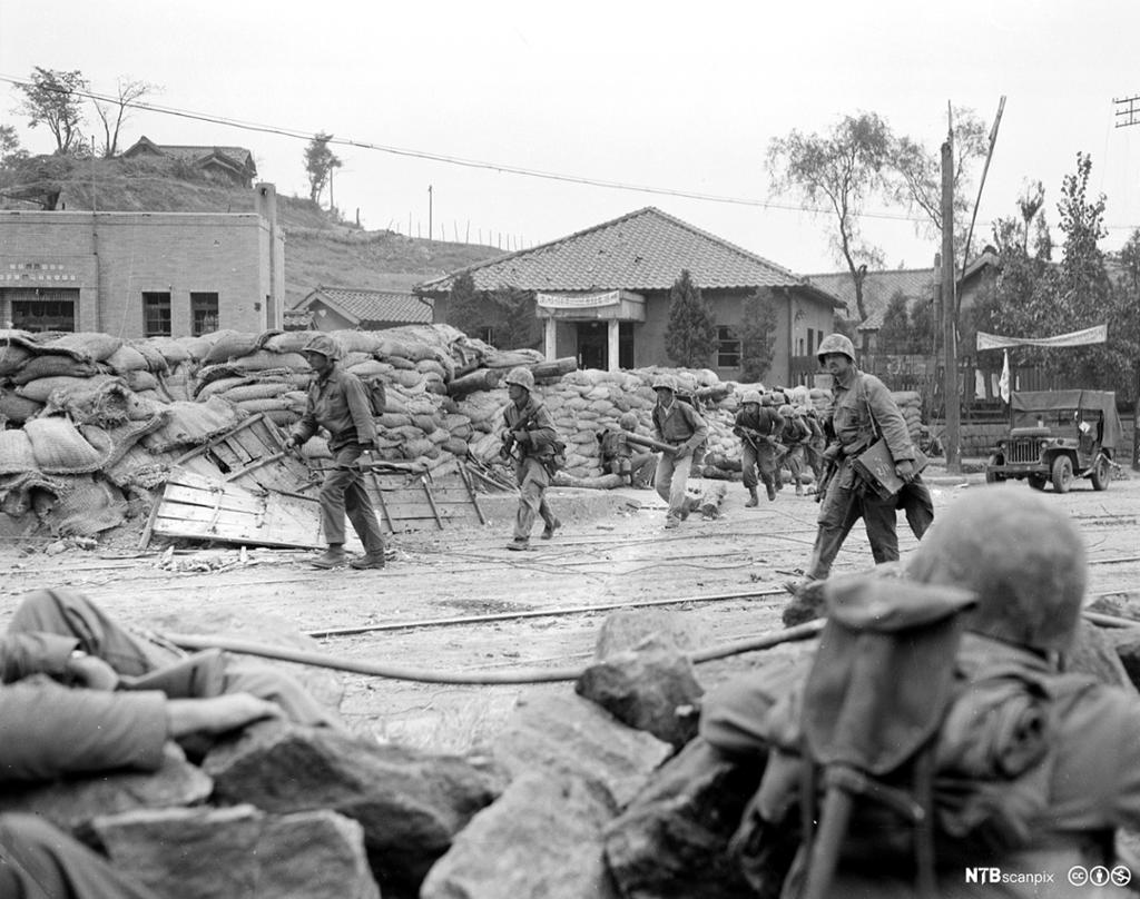 Soldatar beveger seg gjennom øydelagde gater som er i ruinar og med blokkeringer langs gatene av sandsekkar og plank.  Svart-kvitt foto. 
