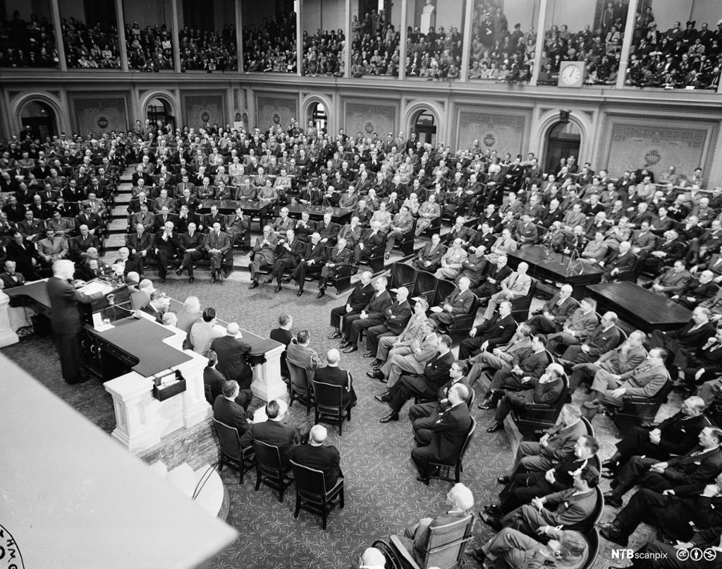 Truman står på en opphøyd talerstol i en stor, halvsirkelformet sal, som er helt full av dresskledde menn. Også på galleriene er det tett i tett med mennesker. Der er det også kvinner. Foto. 