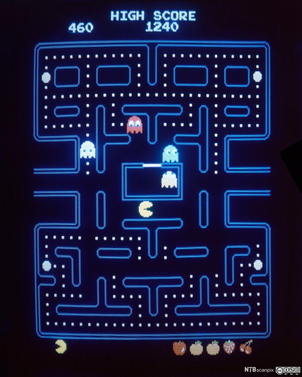 Spillet Pac-Man blir spilt på en gammeldags skjerm. I spillet styrer du Pac-Man, en rund, gul figur med munn, i en labyrint hvor han spiser hvite prikker og frukt/bær, men må passe seg for spøkelsene. Bilde.