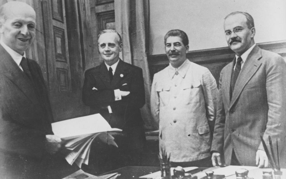 Fire smilande, middelaldrande menn bak eit arbeidsbord. Ein bunke papir blir halden fram. Stalin er litt annleis kledd med ei uniformsliknande jakke. Foto. 