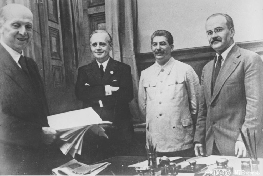 Fire smilende, middelaldrende menn bak et arbeidsbord. En bunke papirer holdes fram. Stalin er litt annerledes kledd med en uniformslignende jakke. Foto. 