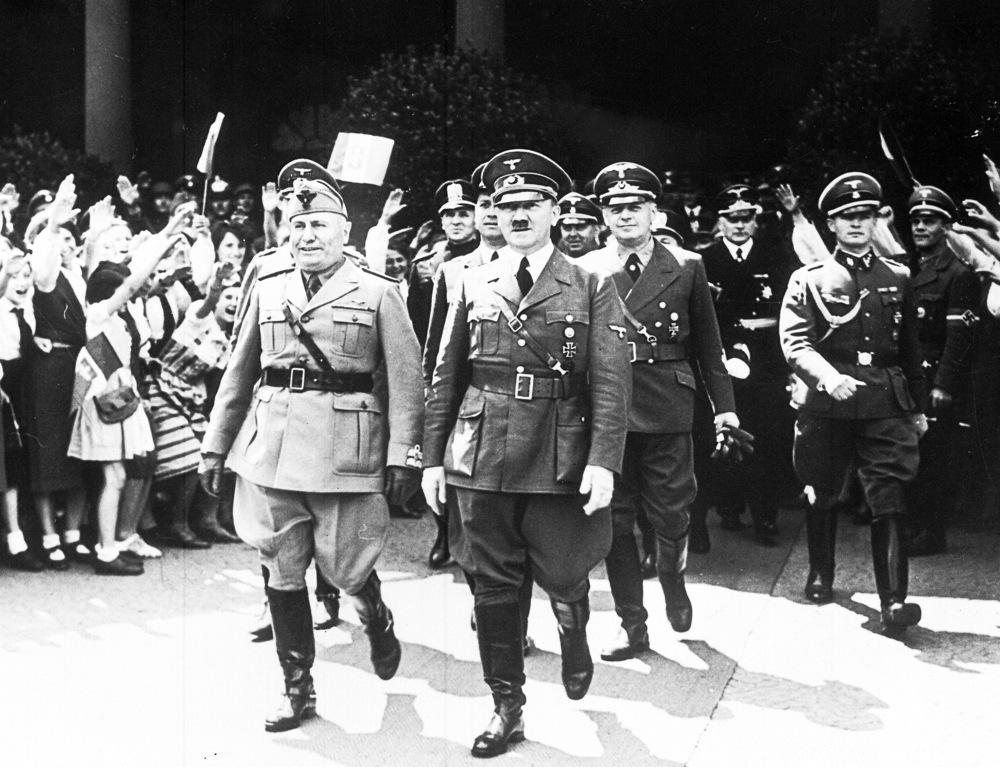 Mussolini og Hitler er iført uniform og går foran flere andre menn i uniform. Det ser ut til å stå en jublende folkemengde på sidene. Foto.