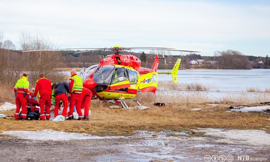 Redningshelikopter ved et islagt vann, ei gruppe redningsmannskaper jobber med en person som ligger på land. Foto.