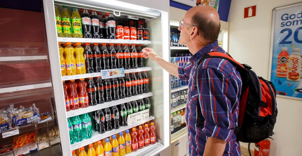 En eldre mann står med utstrakt hånd og skal velge seg en drikkeflaske fra en kjølehylle i en butikk. Foto.