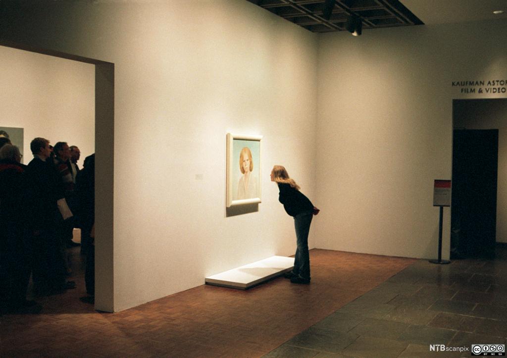 Ei kvinne står åleine i eit galleri-rom og bøyer seg mot eit opplyst måla portrett av ei blond kvinne mens ei folkemengde i neste rom lyttar til ein førelesar. Foto.