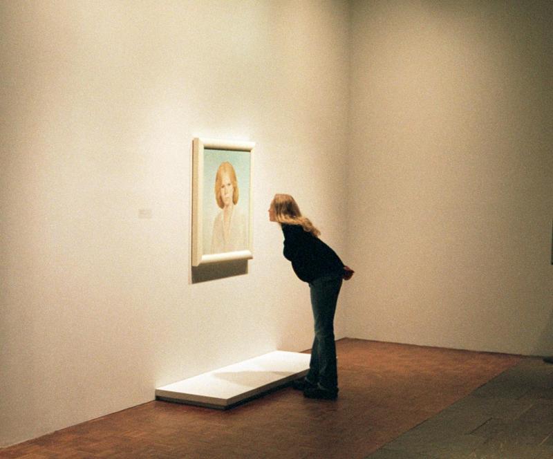 Blond gravid kvinne står alene i et museumsrom og bøyer seg mot et opplyst malt portrett av en blond dame mens en folkemengde i neste rom lytter til en foreleser. Foto.