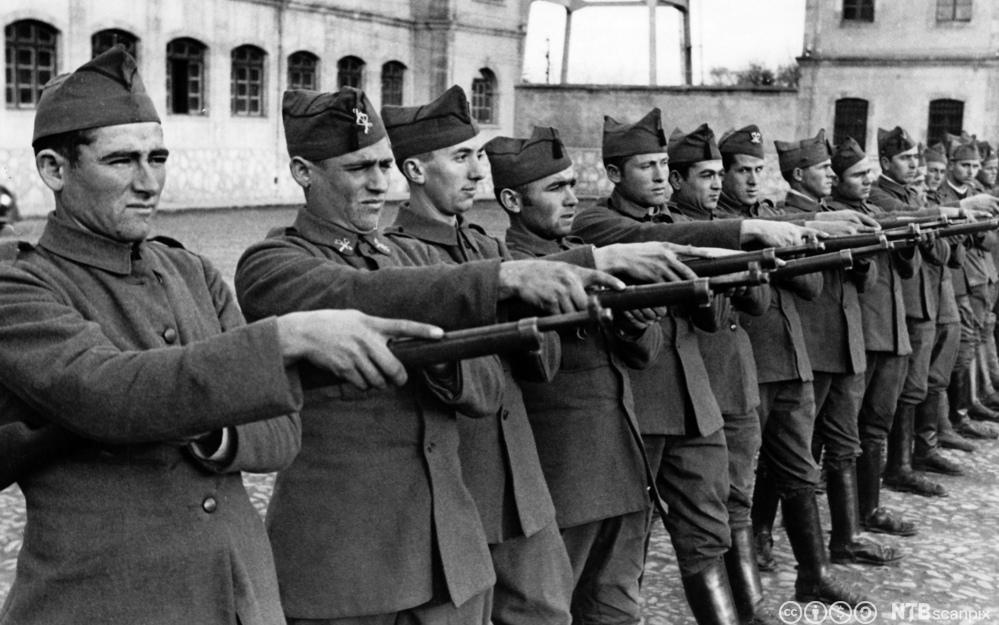 Ei lang rekke uniformskledde menn står oppstilt med geværer rettet framover. Foto. 