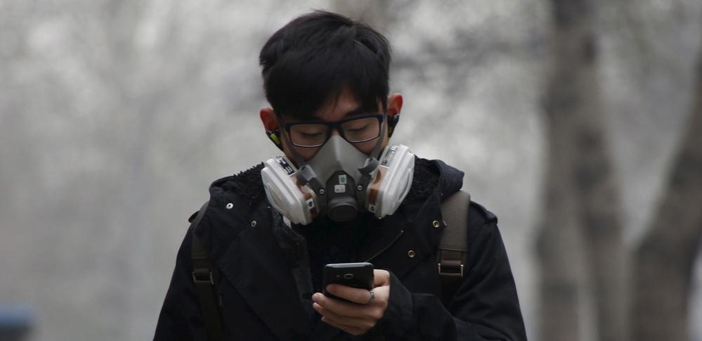 En mann har på seg ei kraftig gassmaske for å beskytte seg mot forurensning. Han holder telefonen sin og ser ned på den. Foto.