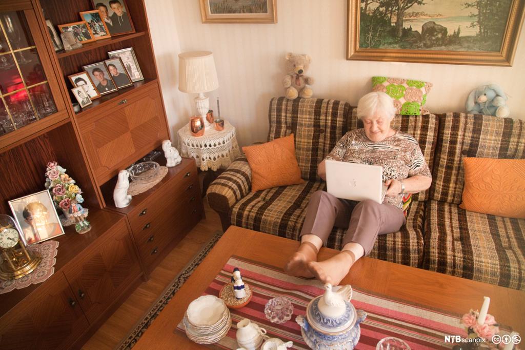 Eldre dame i sofa med laptop på fanget. Foto.