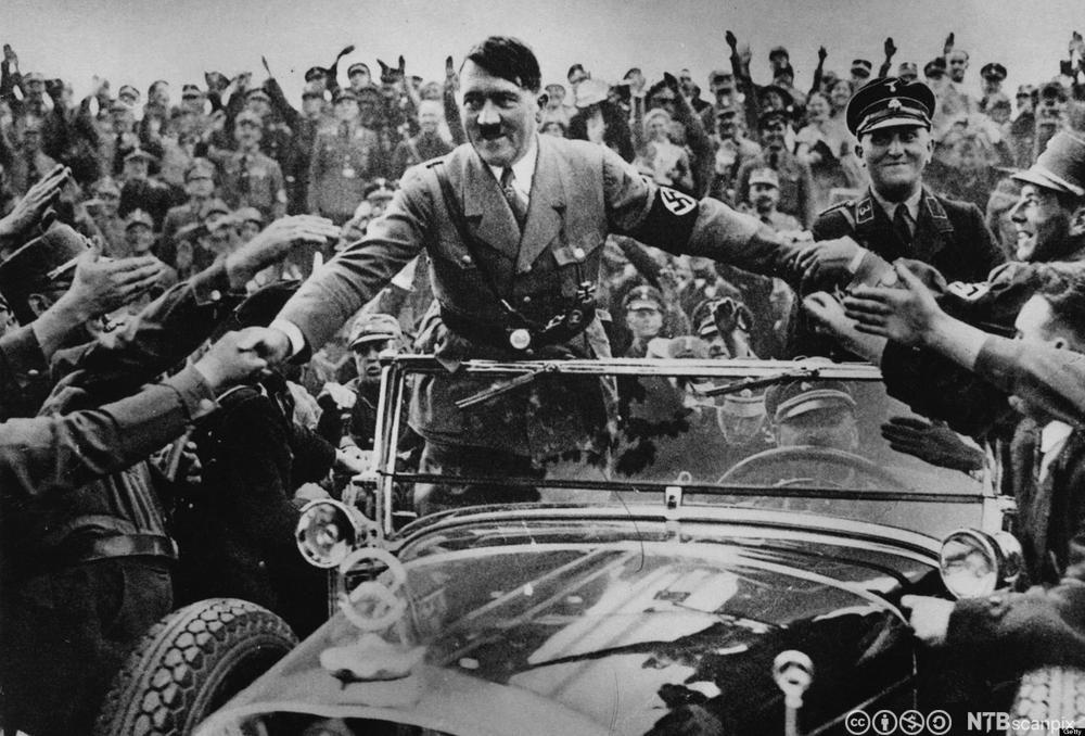 Hitler i uniform og med armbind med hakekors står i ein open bil og helser på tilhengarar langs vegen. Foto.